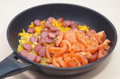 Добавить в сковороду к овощам нарезанные охотничьи колбаски и нарезанные помидоры. Готовить 3-4 минуты.